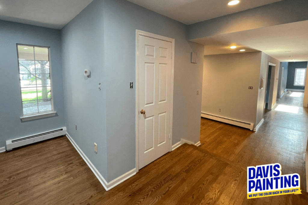 A freshly painted interior hallway with light blue walls and wooden floors. A white door is centered on one wall. Natural light comes through a window on the left. A logo reading "Davis Painting" is in the bottom right corner.