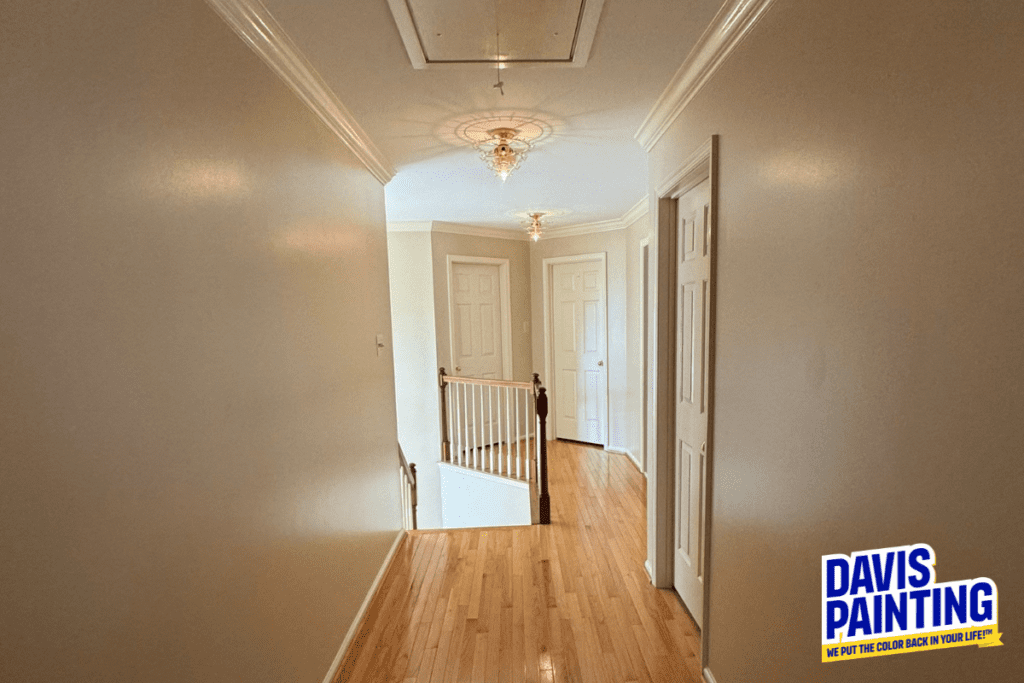 A well-lit hallway with light wood flooring, cream-colored walls, and a decorative light fixture on the ceiling. A staircase with a white railing leads downward. Two closed doors are on the right. A "Davis Painting" logo is visible in the corner.