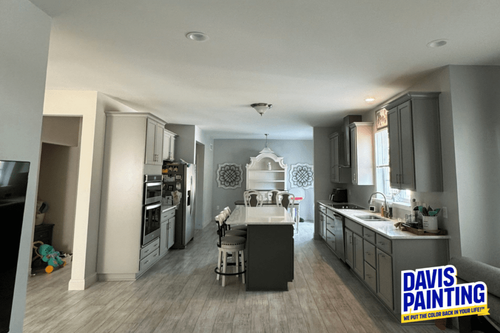 Modern kitchen with gray cabinets, a central island with stools, and stainless steel appliances. Light wood flooring and a dining area with a hutch are visible. A "Davis Painting" logo is in the bottom right corner.