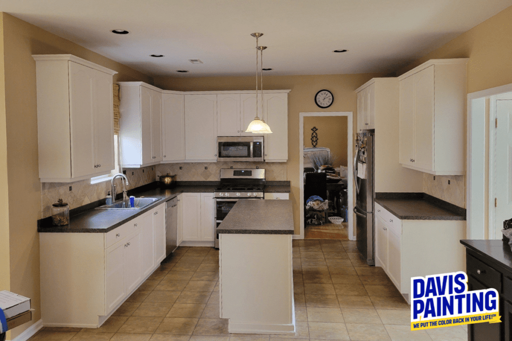 A spacious kitchen with white cabinets, dark countertops, and stainless steel appliances. There's a central island, tiled floor, wall clock, and a ceiling light. The logo in the corner reads "Davis Painting" with the tagline below.