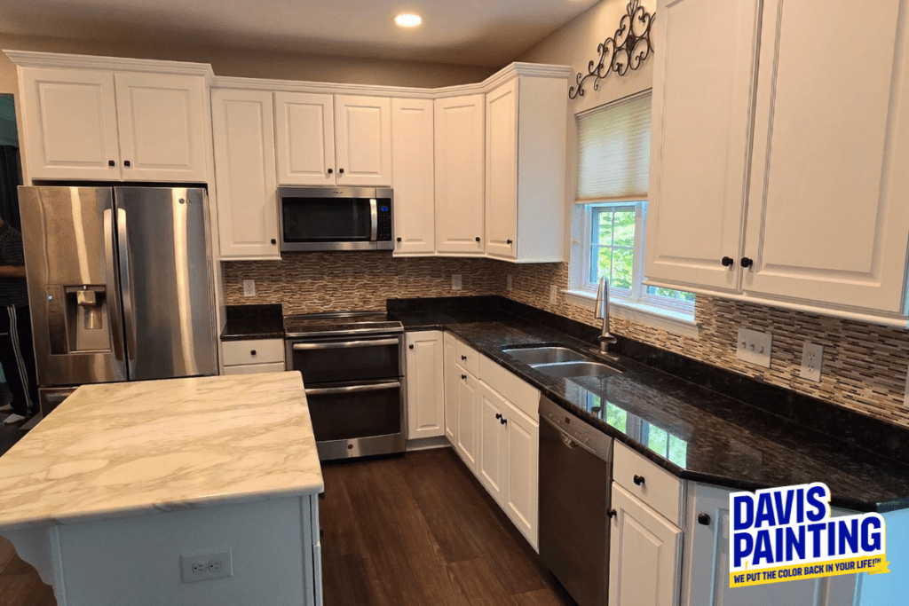 A modern kitchen with white cabinets, stainless steel appliances, a marble island, and dark granite countertops. The backsplash is a mosaic tile design. A window with a blind is above the sink. The Davis Painting logo is in the bottom right corner.