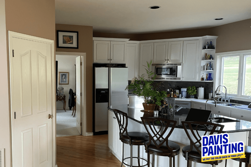 Modern kitchen with white cabinets, stainless steel appliances, and a central island with bar stools. A potted plant sits on the counter. Sunlight streams through a window. A sign in the image reads "Davis Painting.