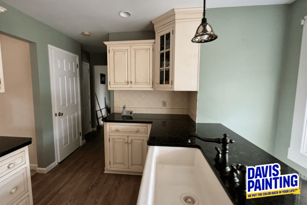 A kitchen with light green walls and cream cabinets. A black countertop, white farmhouse sink, and pendant light are visible. The floor is wood, and a door is on the left. A "Davis Painting" logo is displayed in the bottom right corner.