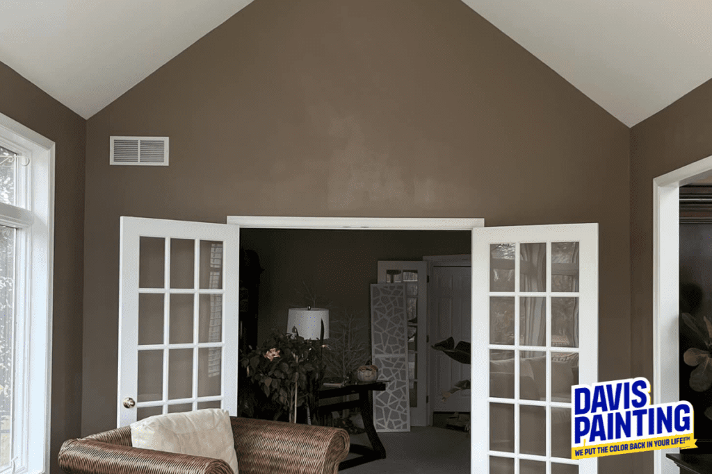 A cozy living room with French doors open to another room. The walls are painted brown, and large windows bring in natural light. A wicker sofa is in the foreground. Plants and a decorative screen are visible inside. "Davis Painting" logo in corner.