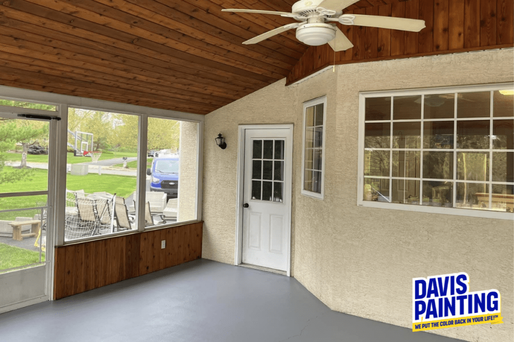 A freshly painted enclosed porch with a wooden ceiling, white fan, and large window. The floor is gray, and the walls are beige. There is a white door leading outside. A "Davis Painting" logo is visible in the corner.