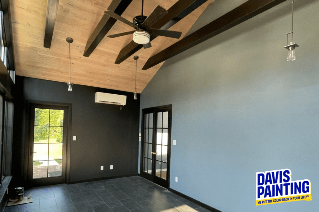 A modern room with a sloped wooden ceiling and exposed beams. The walls are painted in dark and light gray tones. It features a ceiling fan, air conditioning unit, and floor-to-ceiling windows. The room has a sign reading "Davis Painting" in the corner.