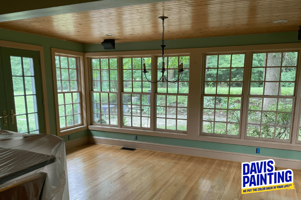 A sunlit room with large windows and a wooden floor. The walls are a soft green, and a chandelier hangs from the wooden ceiling. A glass double door leads outside. The Davis Painting logo is in the bottom right corner.