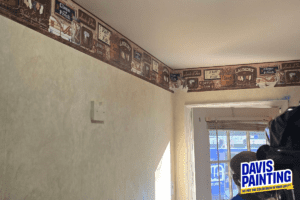 A room with a beige wall and a patterned border near the ceiling, featuring old-fashioned laundry-themed designs. Natural light enters through a window with white blinds. With the "Davis Painting" logo at the bottom right corner, it reminds us that fall is the perfect time to paint your home.