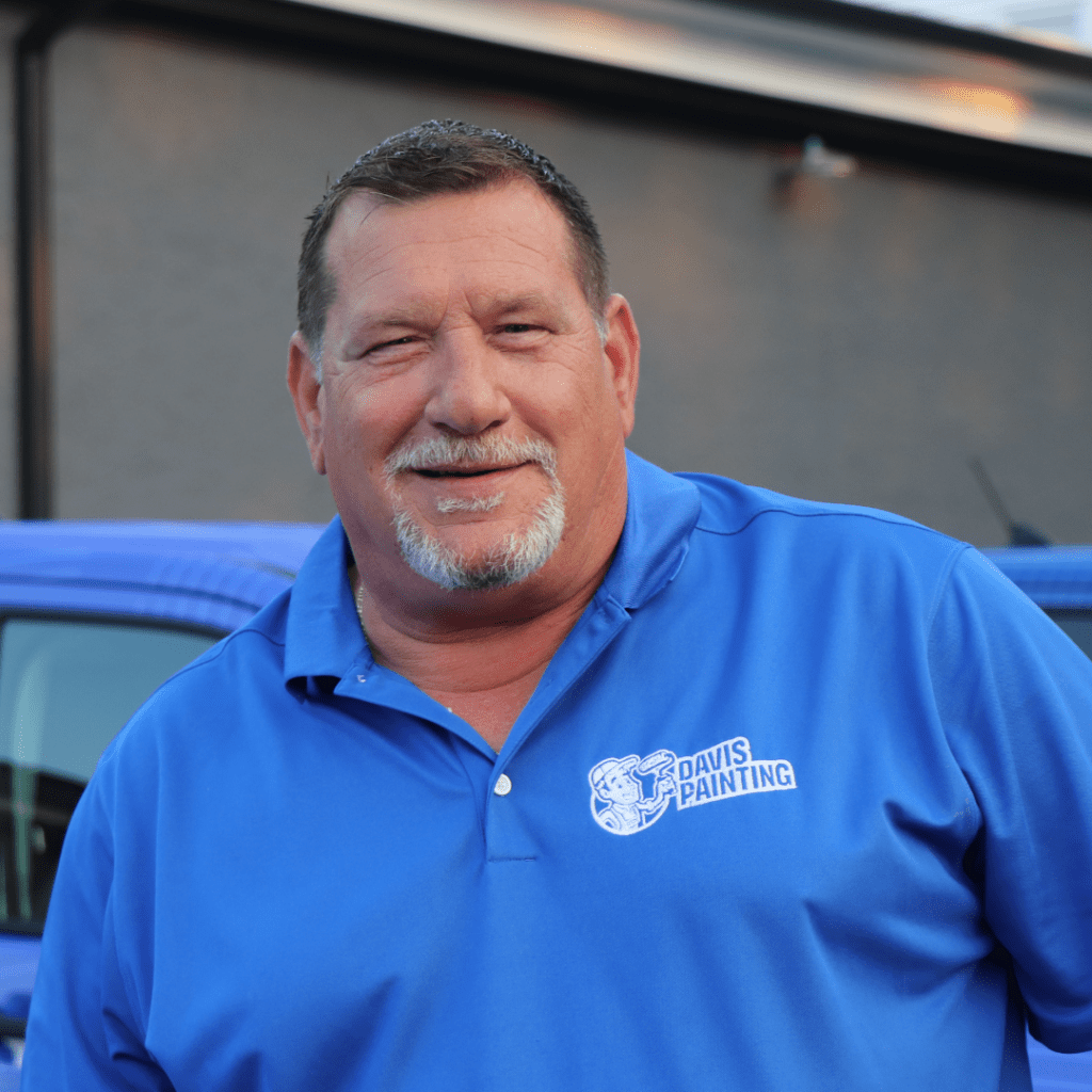 A man in a blue "Davis Painting" polo stands smiling before a blue vehicle, reminiscent of the breezy charm of the Jersey Shore. In the background, a building with a gray wall adds an urban touch.