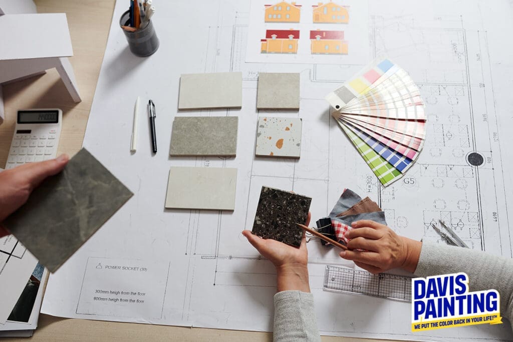 A table with architectural blueprints, interior design samples of tiles, and a color palette. Two people are immersed in selecting materials. A calculator and a Davis Painting logo add to the scene's professional focus.