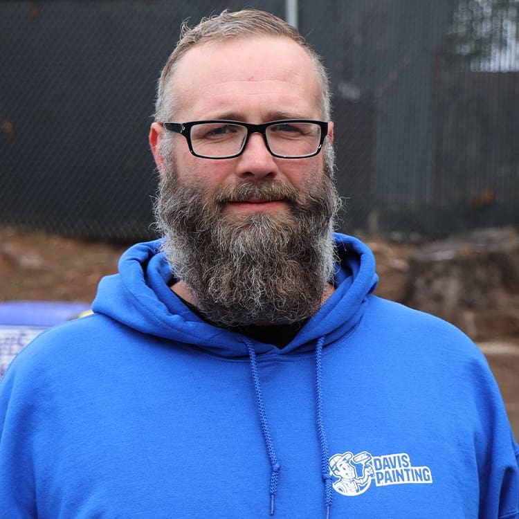 A man wearing glasses and a blue hoodie with the "Davis Painting" logo stands outdoors, reminiscent of a breezy Jersey Shore day. He sports a full beard and short hair against a backdrop of a dark fence and some blurred elements.