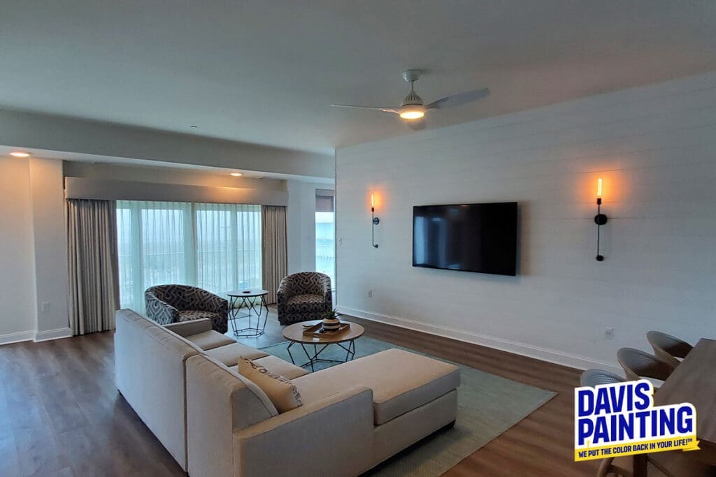 Spacious living room with large windows, light wood flooring, a beige sectional sofa, and patterned armchairs. A flat-screen TV is mounted on a white wall with modern sconces. A ceiling fan is above. The "Davis Painting" logo is in the corner.