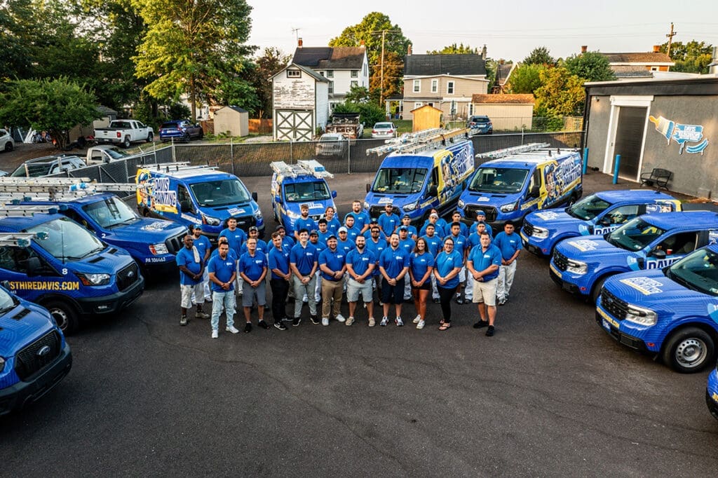 A group of people in blue shirts and beige pants stand in a parking lot, forming a semi-circle in front of several blue service vans with ladders on top. The yellow-and-white branded vans hint at their cabinet painting services. Trees and houses create a serene backdrop.
