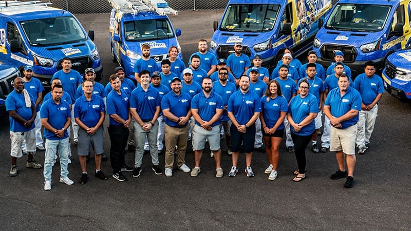 A group of about 25 people wearing blue company shirts stand together outdoors in front of several branded service vans, posing for a team photo in a parking lot—showcasing team spirit and exciting careers opportunities.