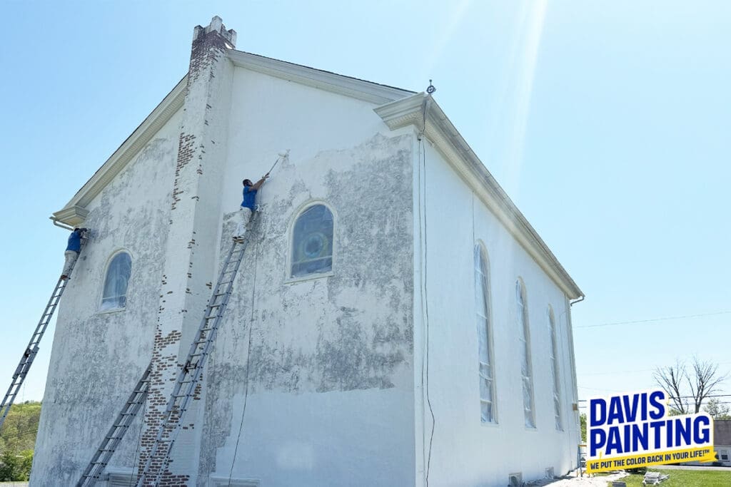 Two painters on ladders work on the exterior wall of a large, old church, undertaking a careful church painting and restoration. The wall is partly weathered and partly freshly painted white. A "Davis Painting" logo appears at the bottom right.