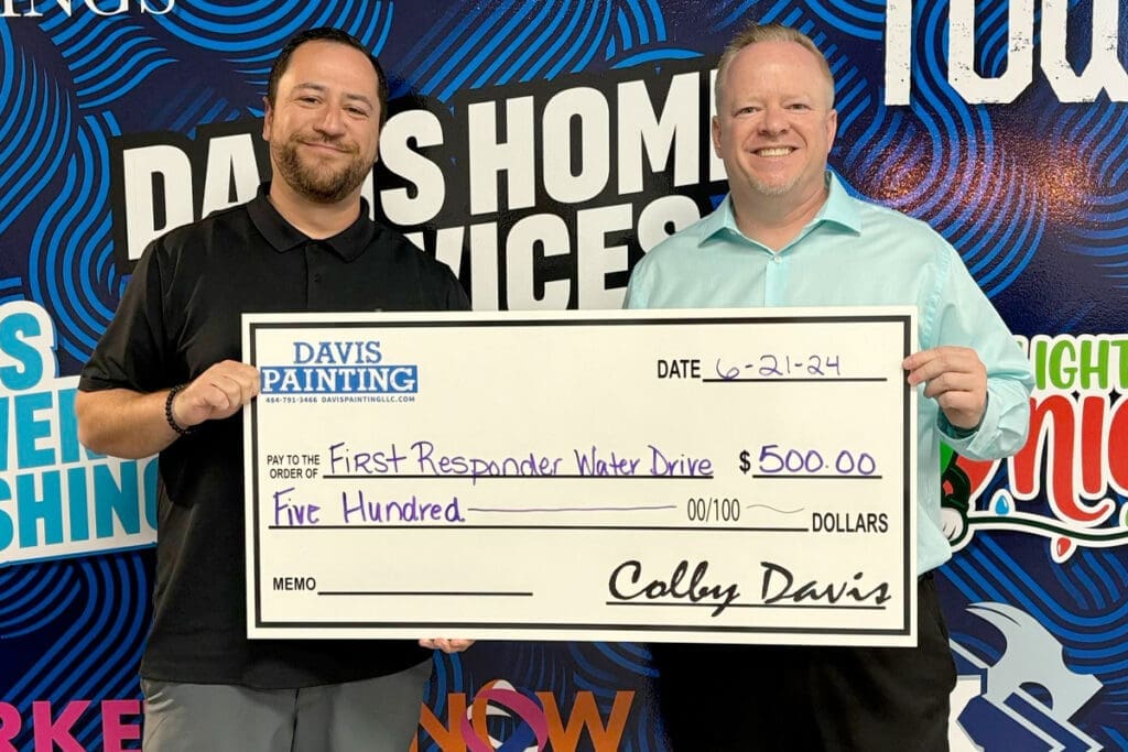 Two smiling men hold a large check for $500 from Davis Painting, a local cabinet painting expert, dated 6-21-24 and made out to the "First Responder Water Drive." Colorful company graphics fill the background.