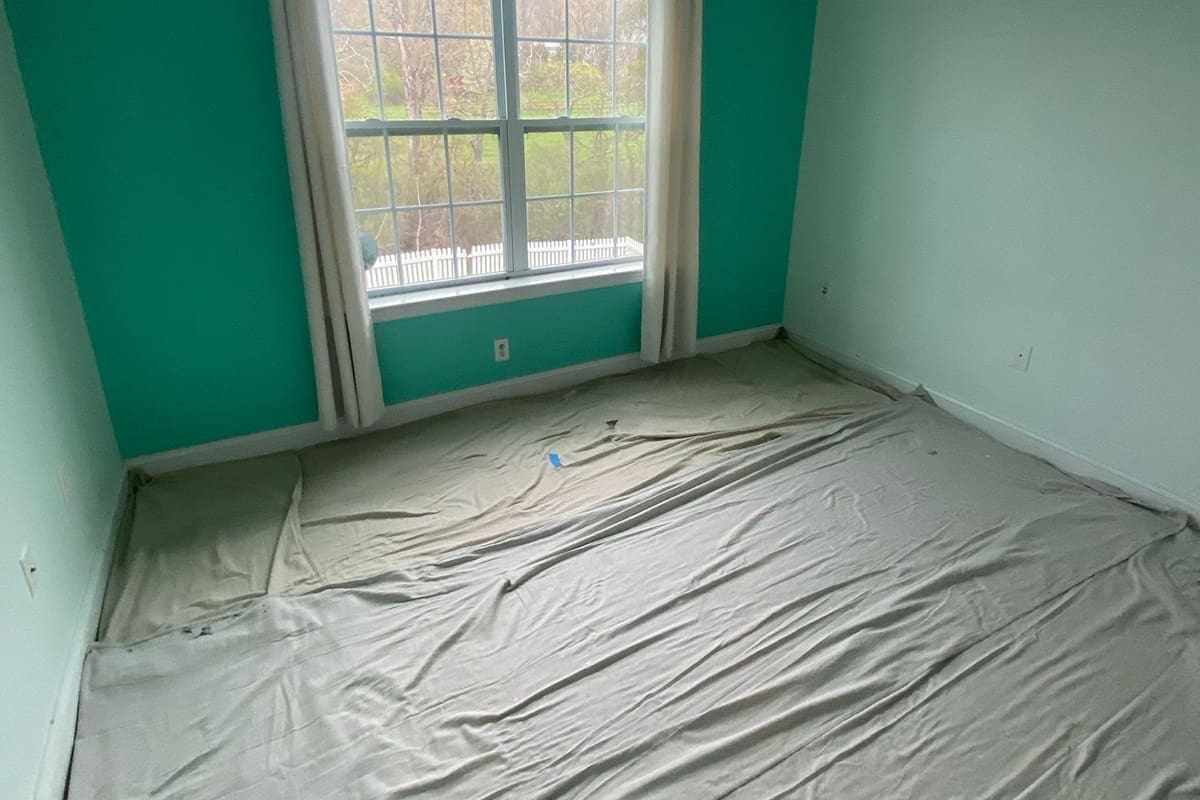 An empty room with teal and light green walls, a large window with white curtains, and floors covered with drop cloths, reflecting current interior painting trends in preparation for a fresh renovation.