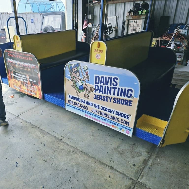 A blue and yellow amusement park ride car on the Wildwoods Boardwalk displays a Davis Painting ad at the Jersey Shore, showing a cartoon man relaxing with a paint roller. A sign on top indicates a $5.00 price.