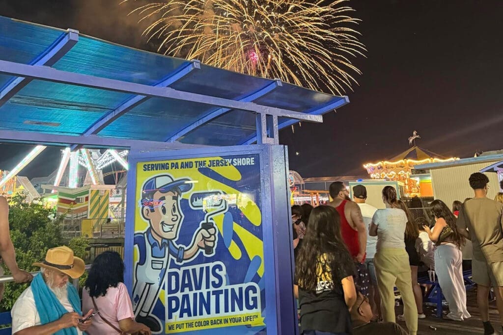 People gather at a lively fairground at night, with colorful fireworks lighting up the sky. In the foreground, a large sign advertises “Davis Painting,” who partners with the Wildwoods Boardwalk to ride alongside the Wildwood Tramcar. Carnival rides and booths glow behind.